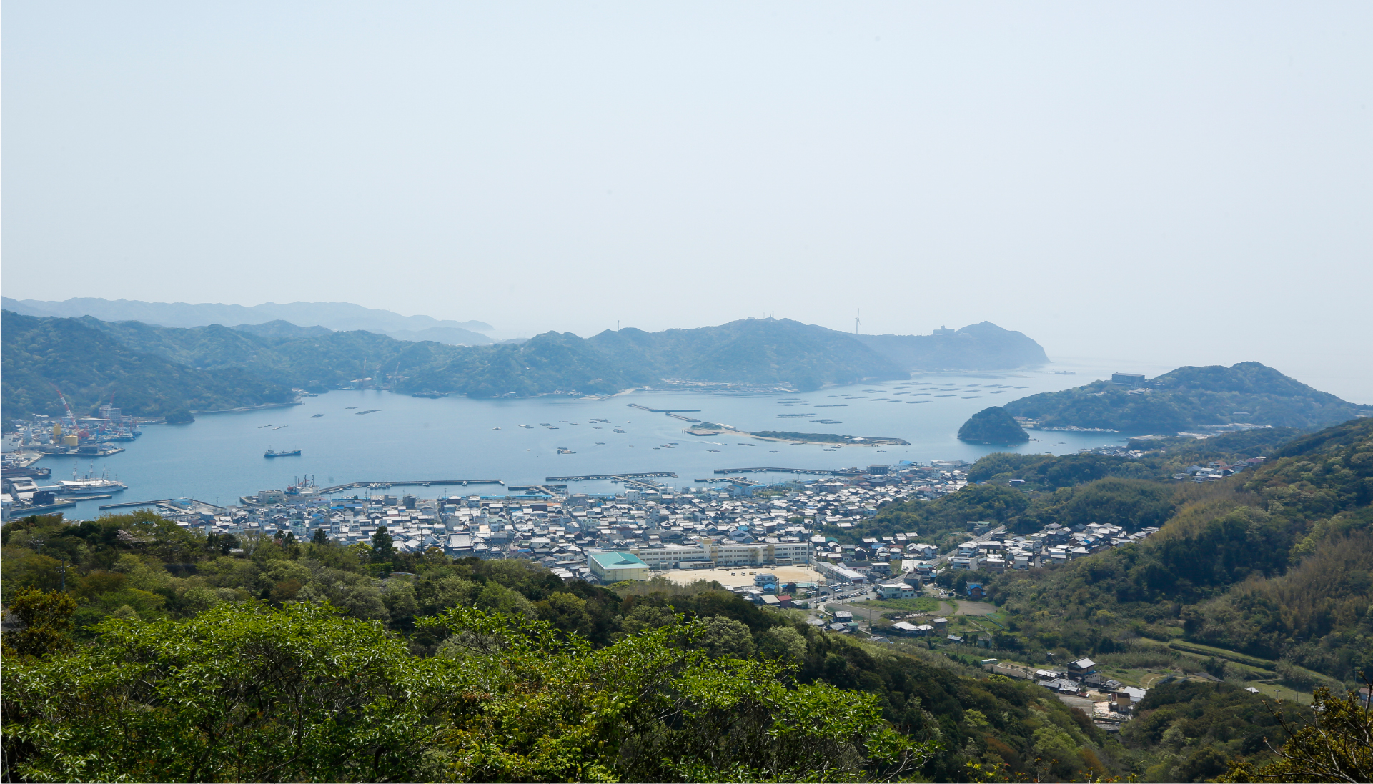Aerial view of Awaji Island's coastal town nestled between forested hills and calm sea, revealing the island's peaceful and unhurried character