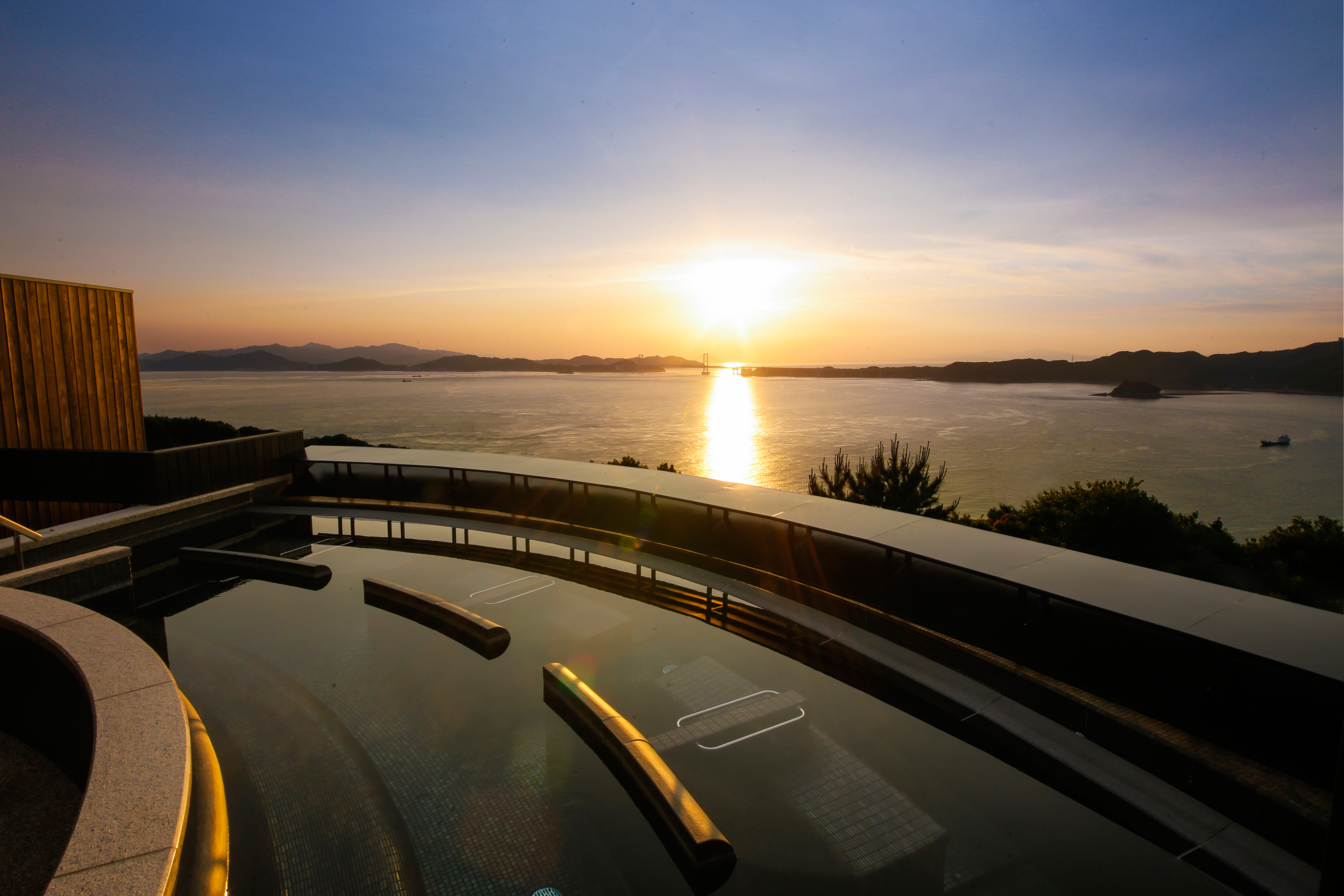Golden sunset over a coastal road beside calm waters and distant mountain silhouettes, capturing the tranquil scenery surrounding Hotel New Awaji Group's destinations