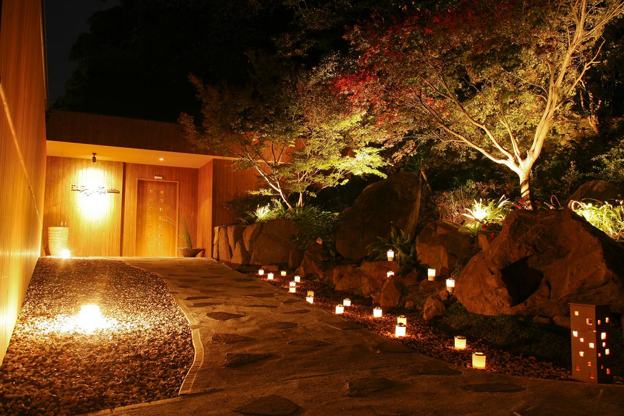 Illuminated autumn maple trees and candlelit stone pathway in a Japanese garden at night, creating an atmosphere of warmth and quiet beauty
