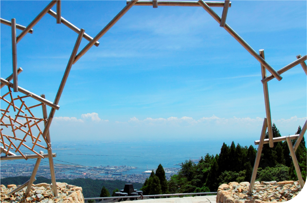 Daytime coastal panorama of Kobe framed through a geometric steel structure, where mountain, sea, and city exist in a rare and quietly confident balance