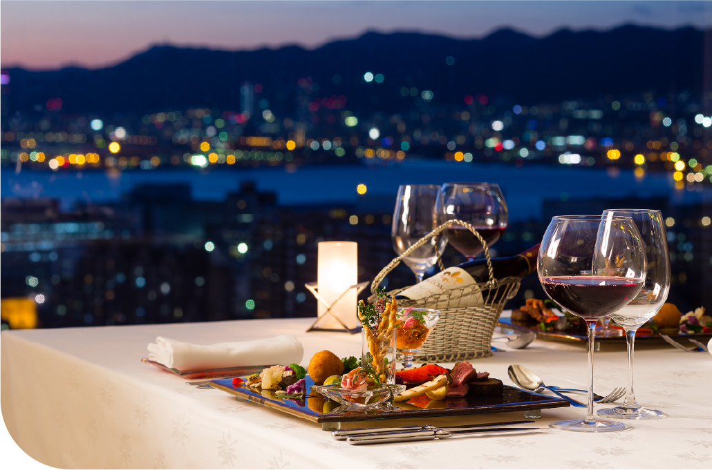 Candlelit dinner table with wine and seasonal dishes set against Kobe's full city light horizon at night, where refined hospitality and the energy of the city meet as one