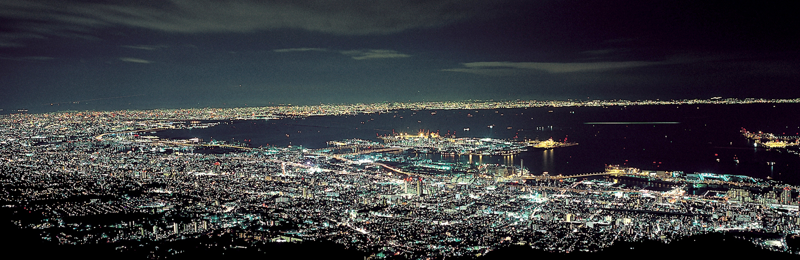 Sweeping panoramic view of Kobe's city lights stretching from mountain foothills to the harbor at night, revealing the full scale and luminous energy of one of Japan's most cosmopolitan coastal cities