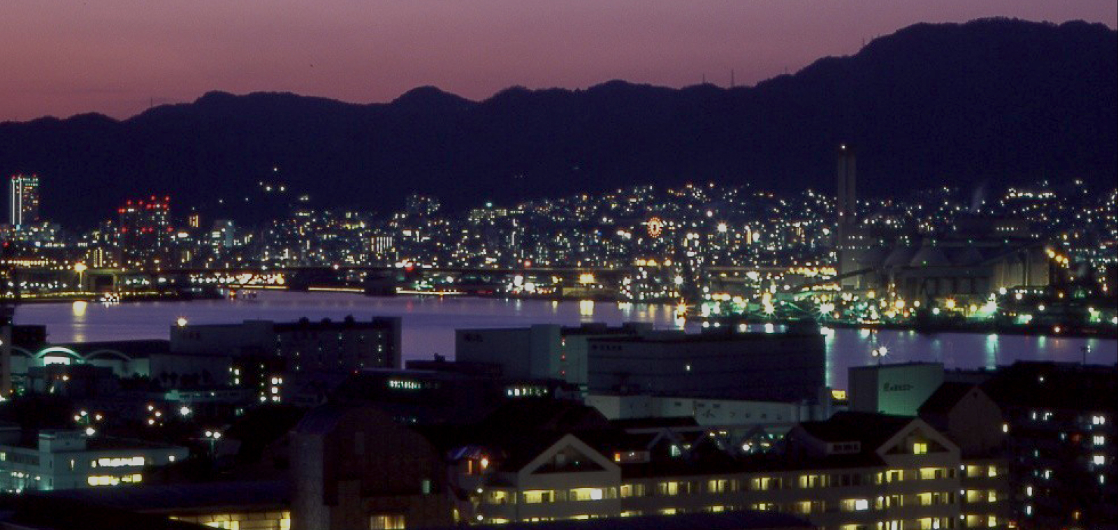 Kobe harbor city lights reflecting on the water at dusk with mountain silhouettes, capturing the vibrant energy and coastal sophistication of the city