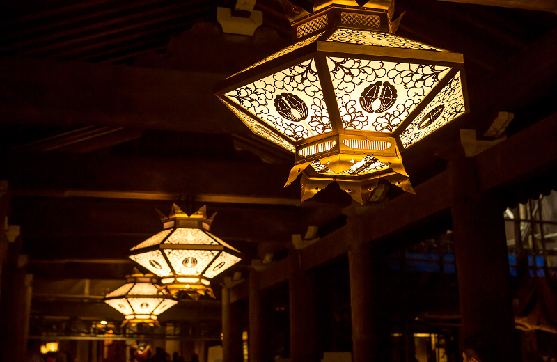 Temple roofline amid autumn maple branches and ornate golden shrine lanterns glowing in a dark timber hall, capturing the layered beauty and sacred atmosphere of Kyoto