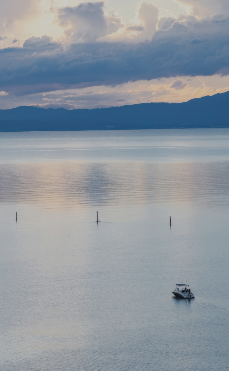 Vast still waters of Lake Biwa reflecting soft clouds at dusk with a lone boat on the horizon, capturing the lake's openness and quiet grandeur