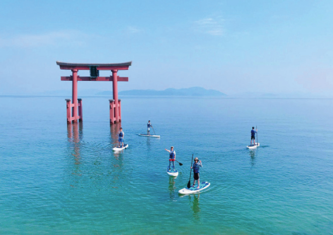Paddleboarders gliding toward a red torii gate on the turquoise waters of Lake Biwa, blending active discovery with cultural wonder