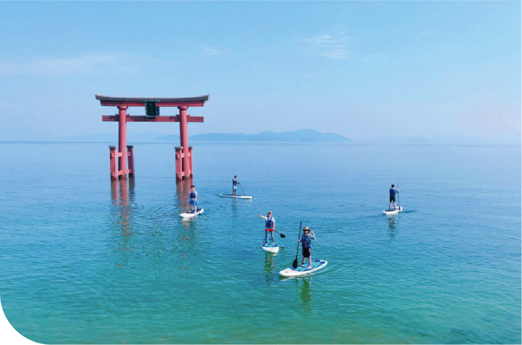 Paddleboarders approaching a traditional red torii gate on the still turquoise waters of Lake Biwa, offering a rare perspective on the lake's sacred shoreline