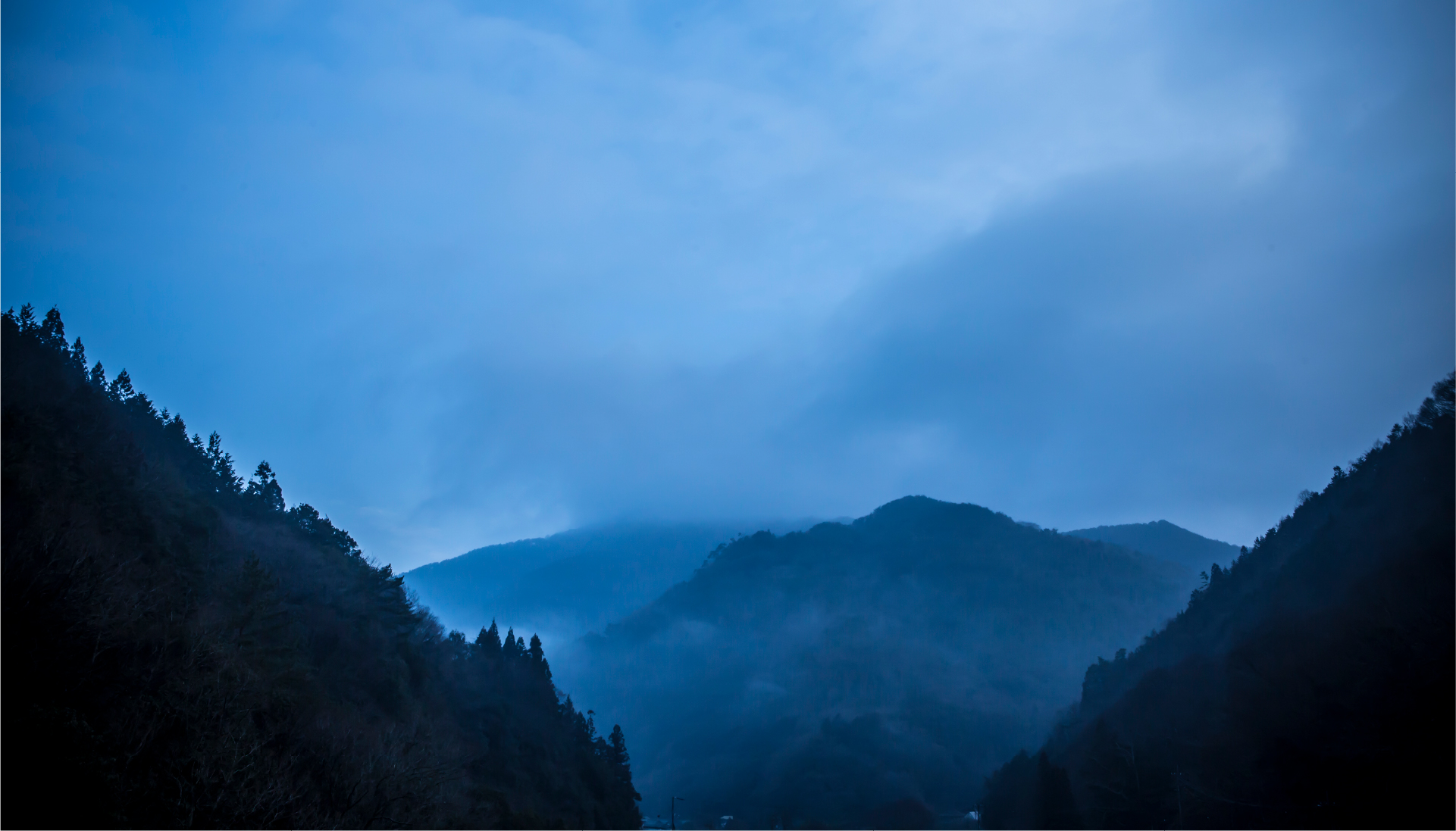Misty mountain valley in Shikoku at twilight, where forested ridgelines dissolve into blue fog and the landscape invites slow, reflective discovery
