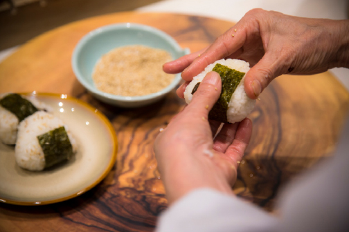Hands carefully wrapping a nori onigiri beside a bowl of miso soup, a moment of simple, artisanal care that defines the culinary spirit of Shikoku and Okayama
