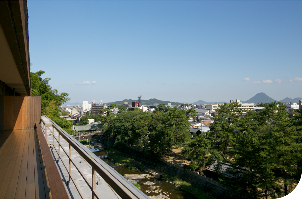 Elevated view over a Shikoku coastal town with mountain and sea stretching to the horizon, capturing the quiet vastness of a region shaped by land and water